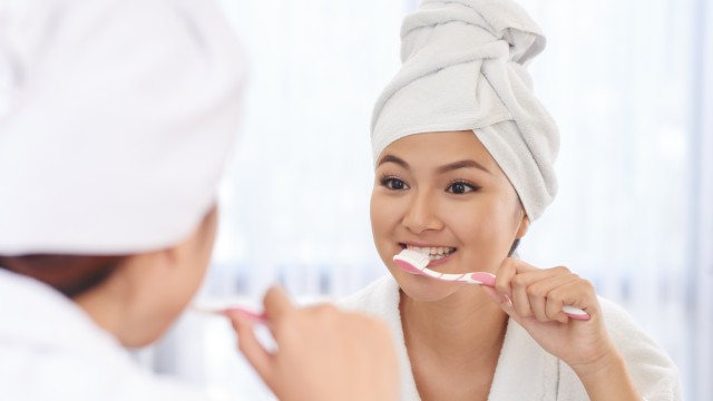 Primer plano de mujeres jóvenes cepillándose los dientes frente al espejo en su bata y con una toalla en la cabeza